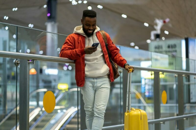 Smiling African American guy using smartphone, checking incoming messages after arrival to airport